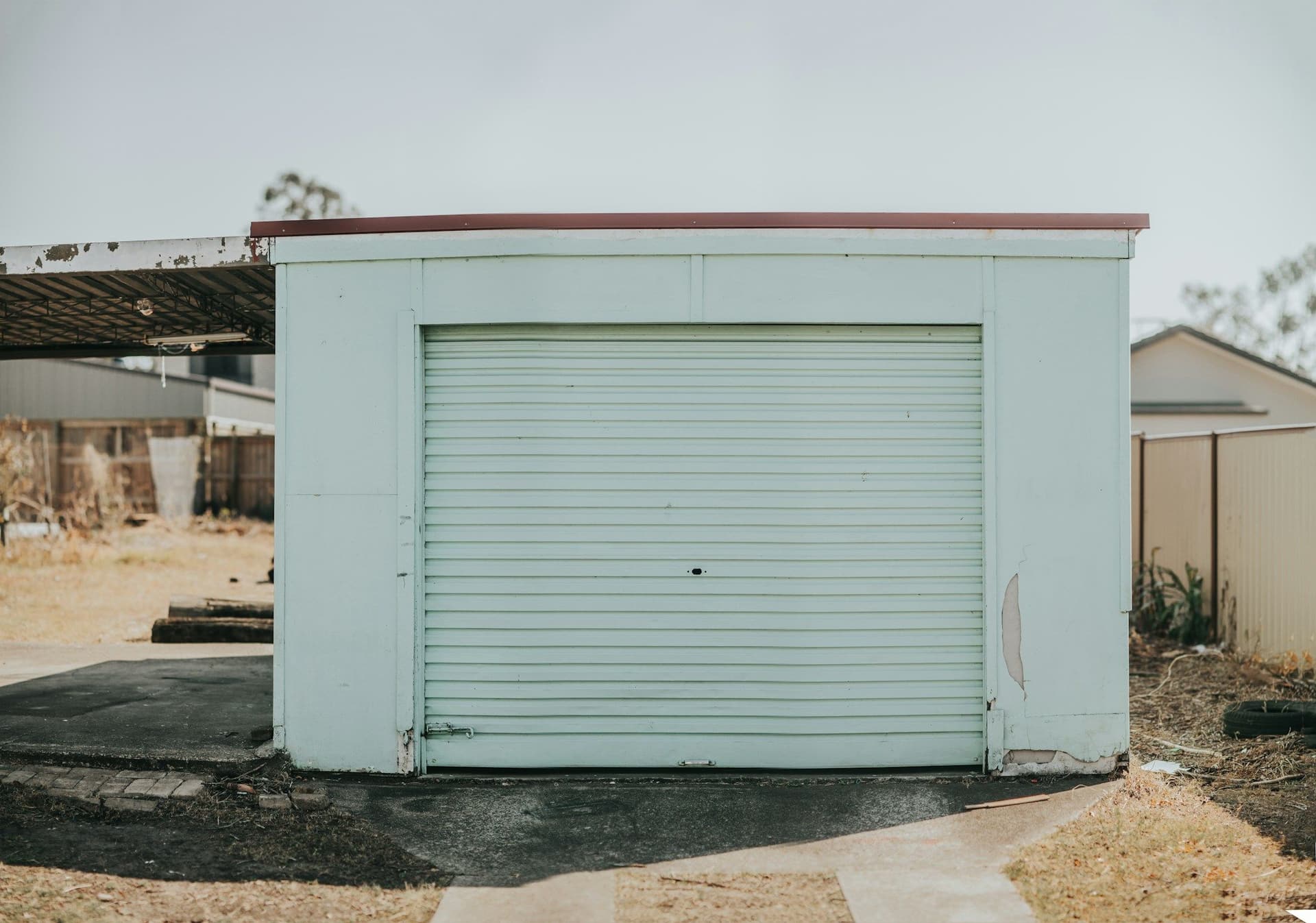 A small garage with a closed pale green roller door standing on dry ground under a pale sky