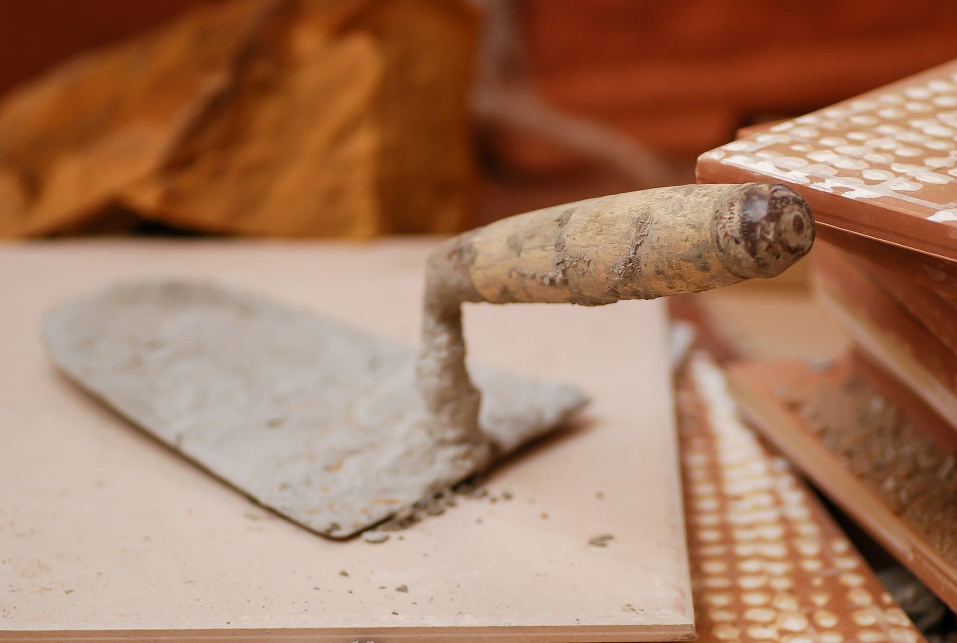 Steel trowel loaded with gray mortar resting on a tile beside a stack of tiles