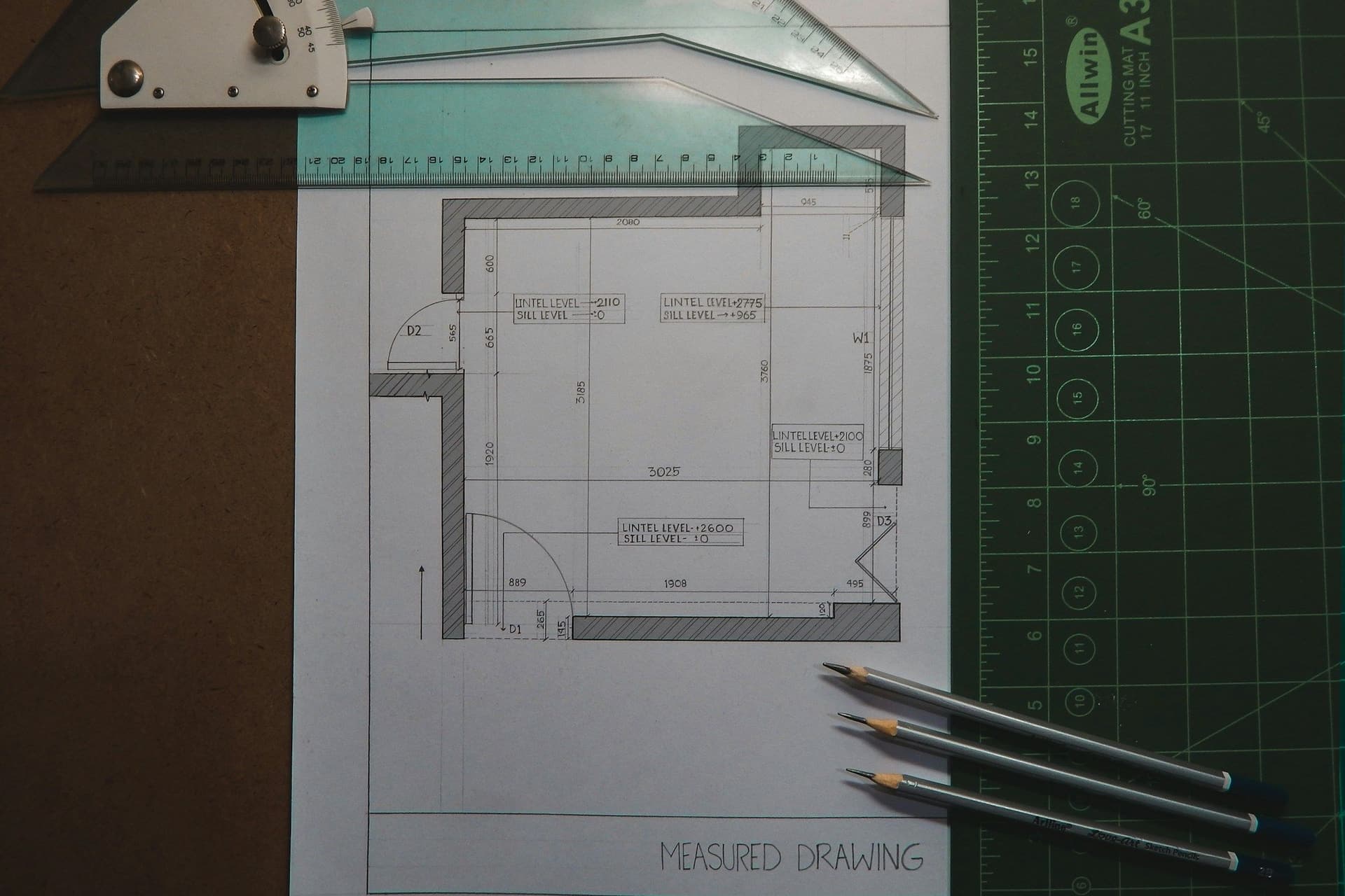 A measured floor plan drawing on a cutting mat with a ruler, a drafting triangle and pencils