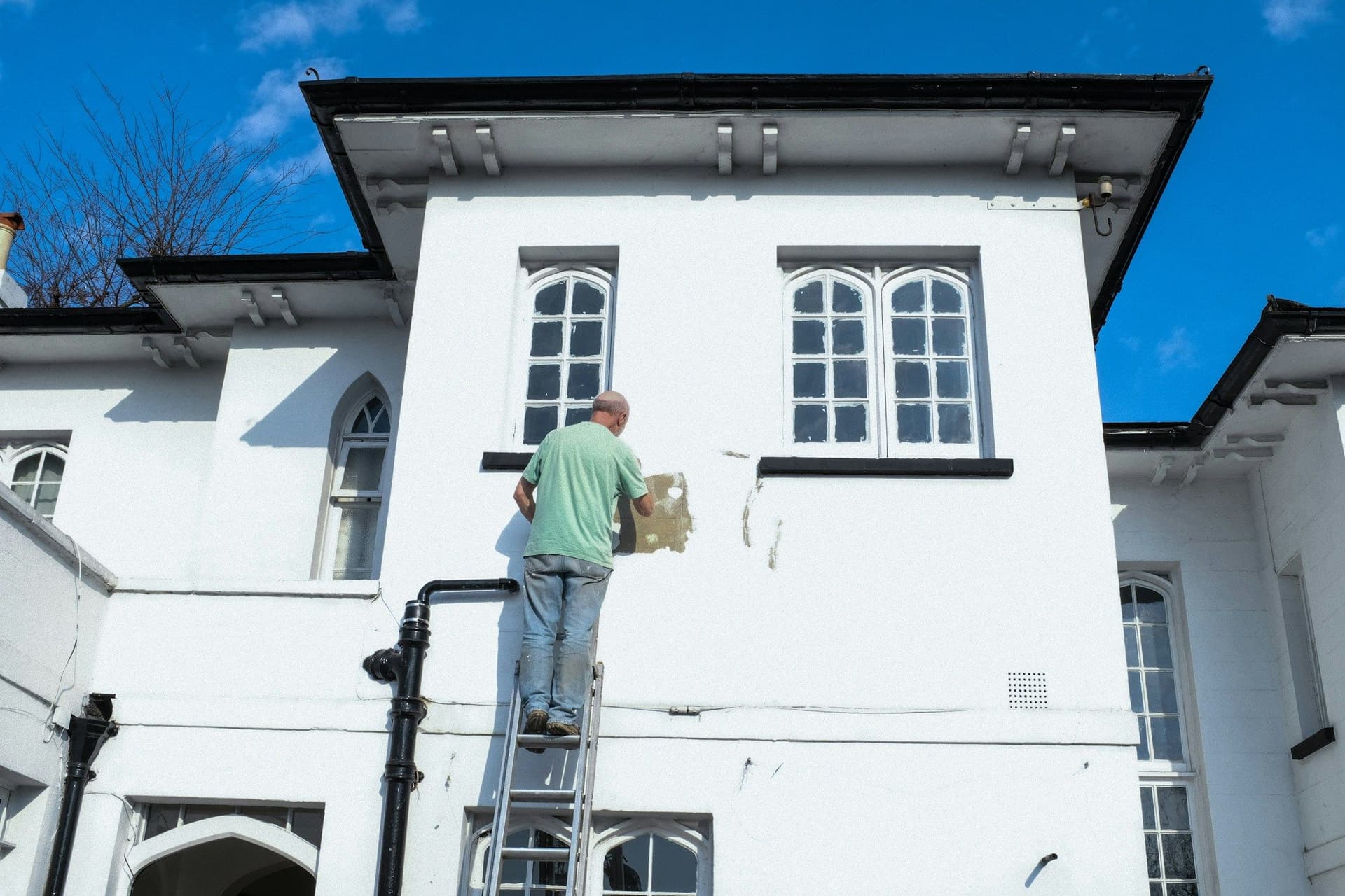 A man on a ladder scraping old paint from the upper wall of a white house under a blue sky