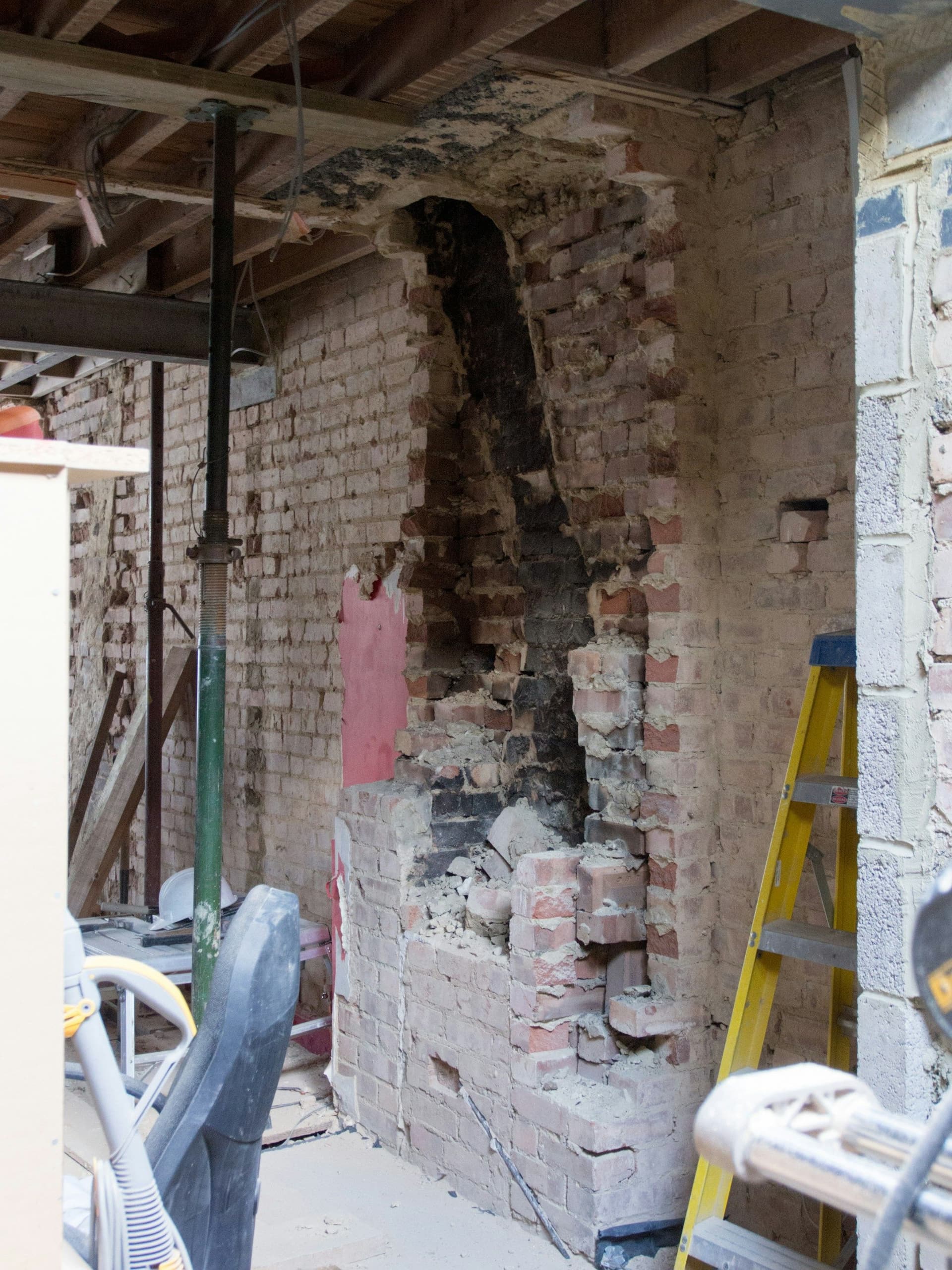 A brick wall partly demolished inside a room, with a yellow ladder, exposed ceiling joists and a steel support