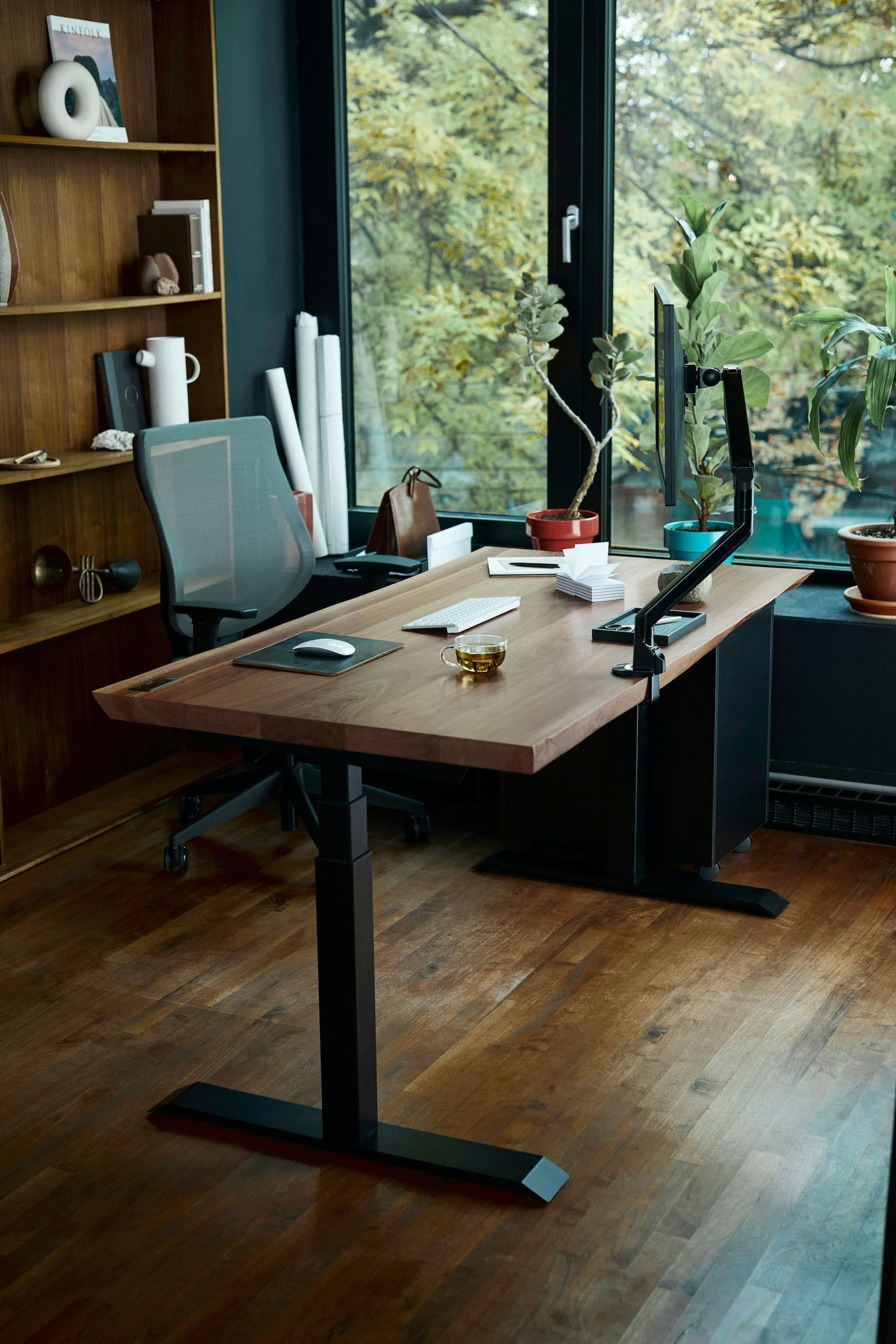 Home office with a wooden standing desk, a mesh chair, a wall of shelves and a large window onto trees