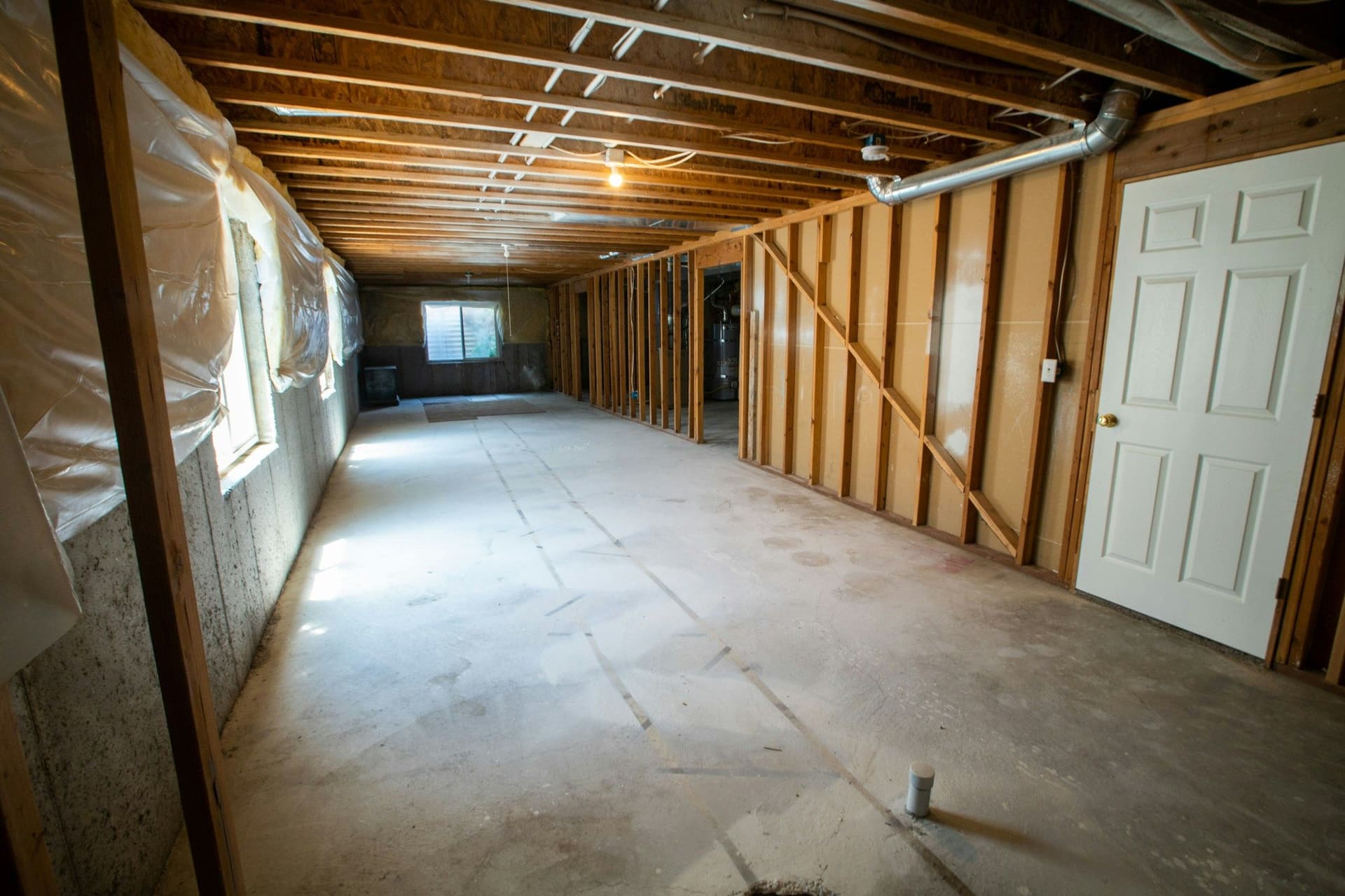 A long basement stripped to its framing, with exposed joists, plastic sheeting on one wall and a white door