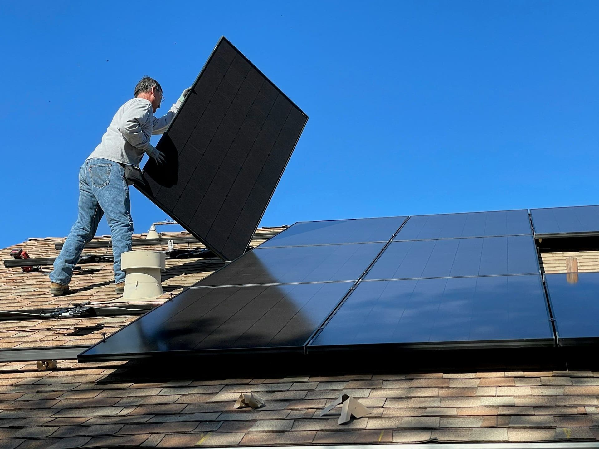 A man carries a black solar panel across a shingled roof under a clear blue sky