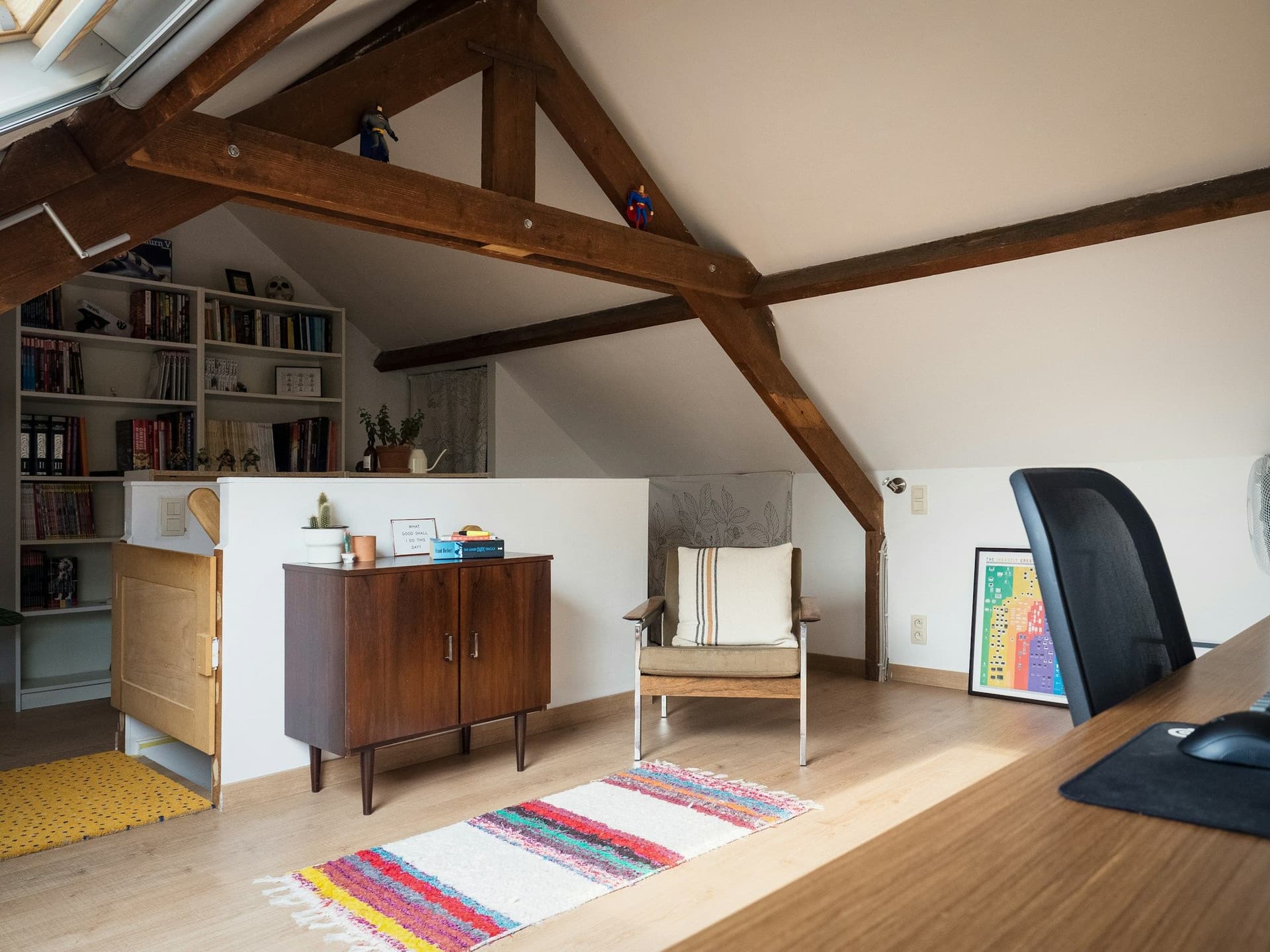 Attic study with exposed timber beams, bookshelves, a desk and a rug on the wood floor