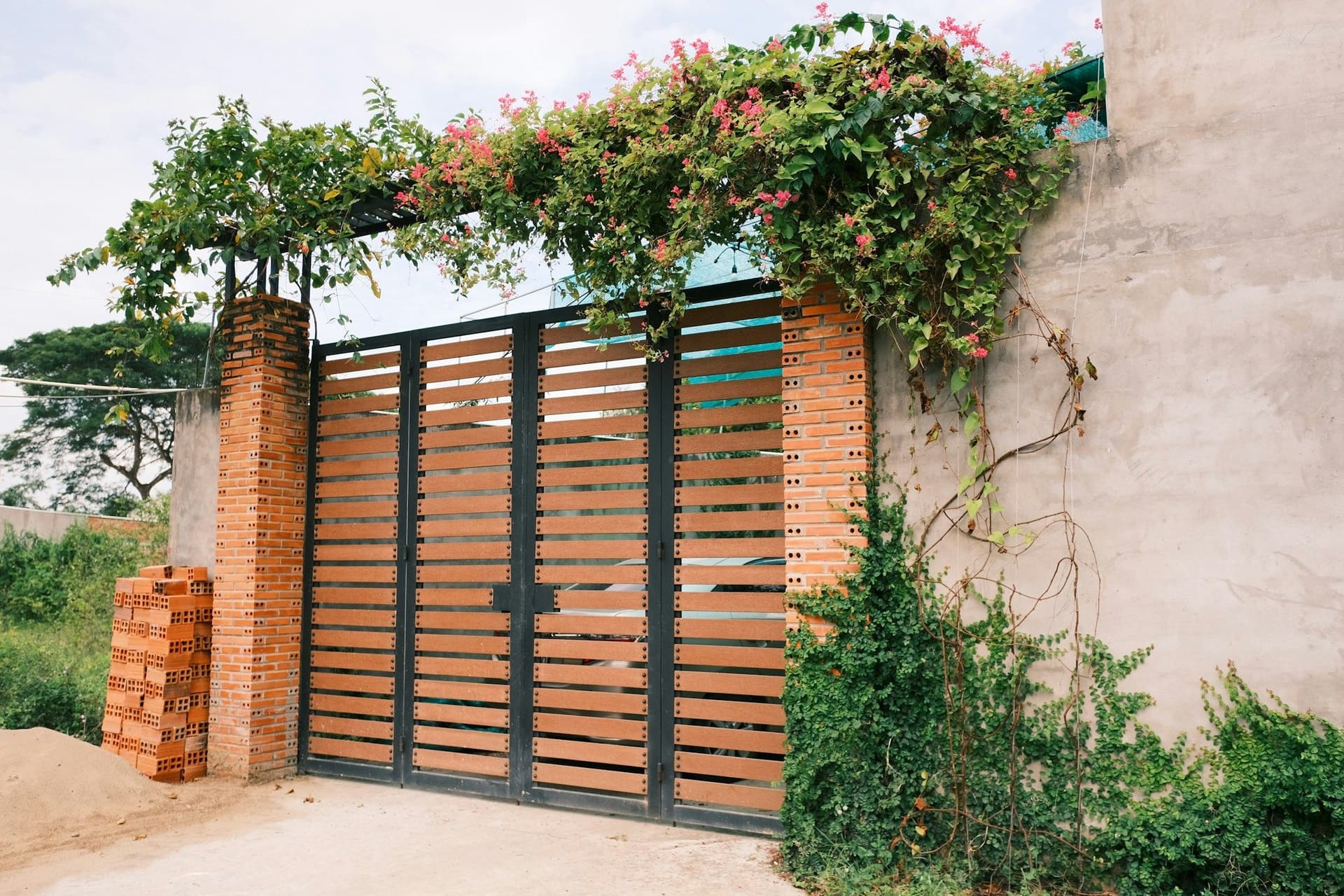 Double gate of wood slats in a dark steel frame between brick pillars under flowering vines
