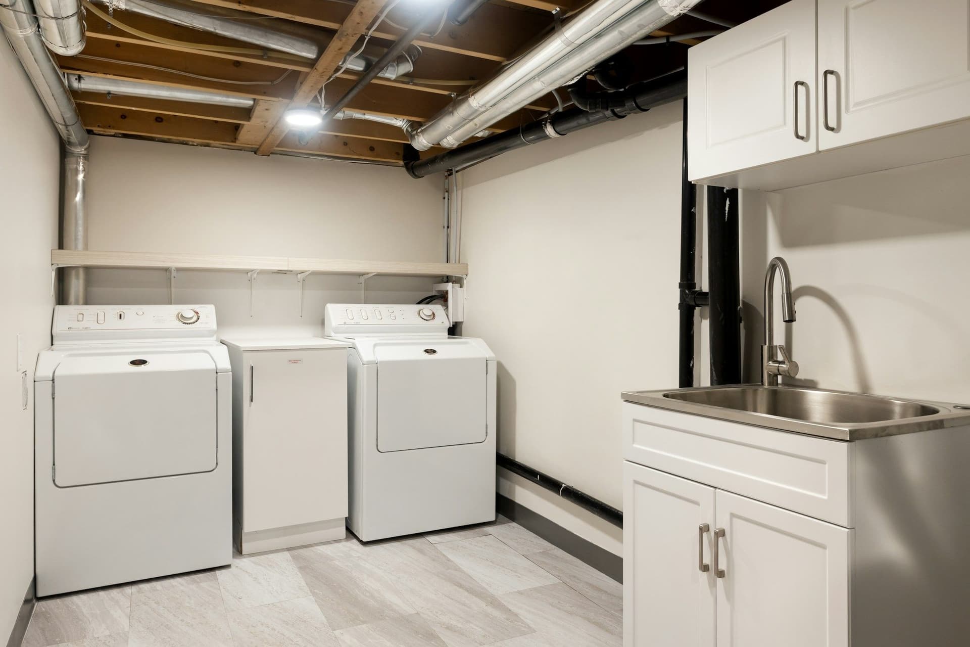 Utility room with a washer and dryers, a sink and white cabinets under exposed pipes and joists