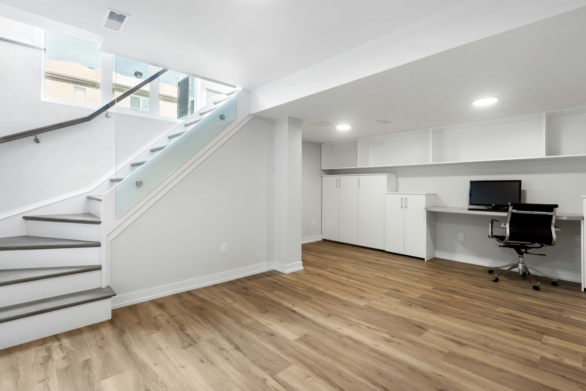 Finished basement with a glass-railed staircase, a wall of white built-in cabinets and a desk with a black chair