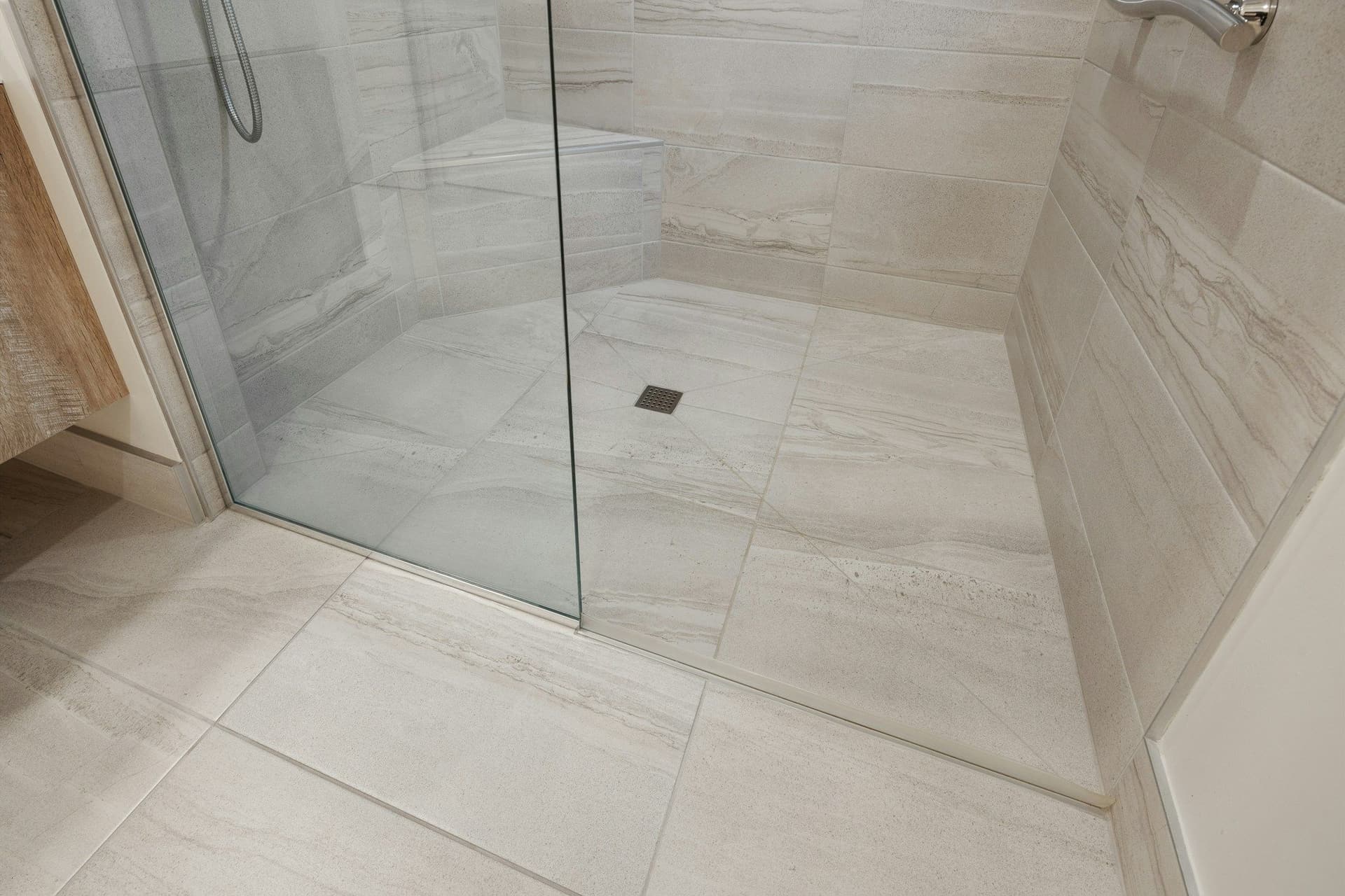 Large pale floor tiles continuing from the bathroom into a shower behind a glass panel, with a small drain