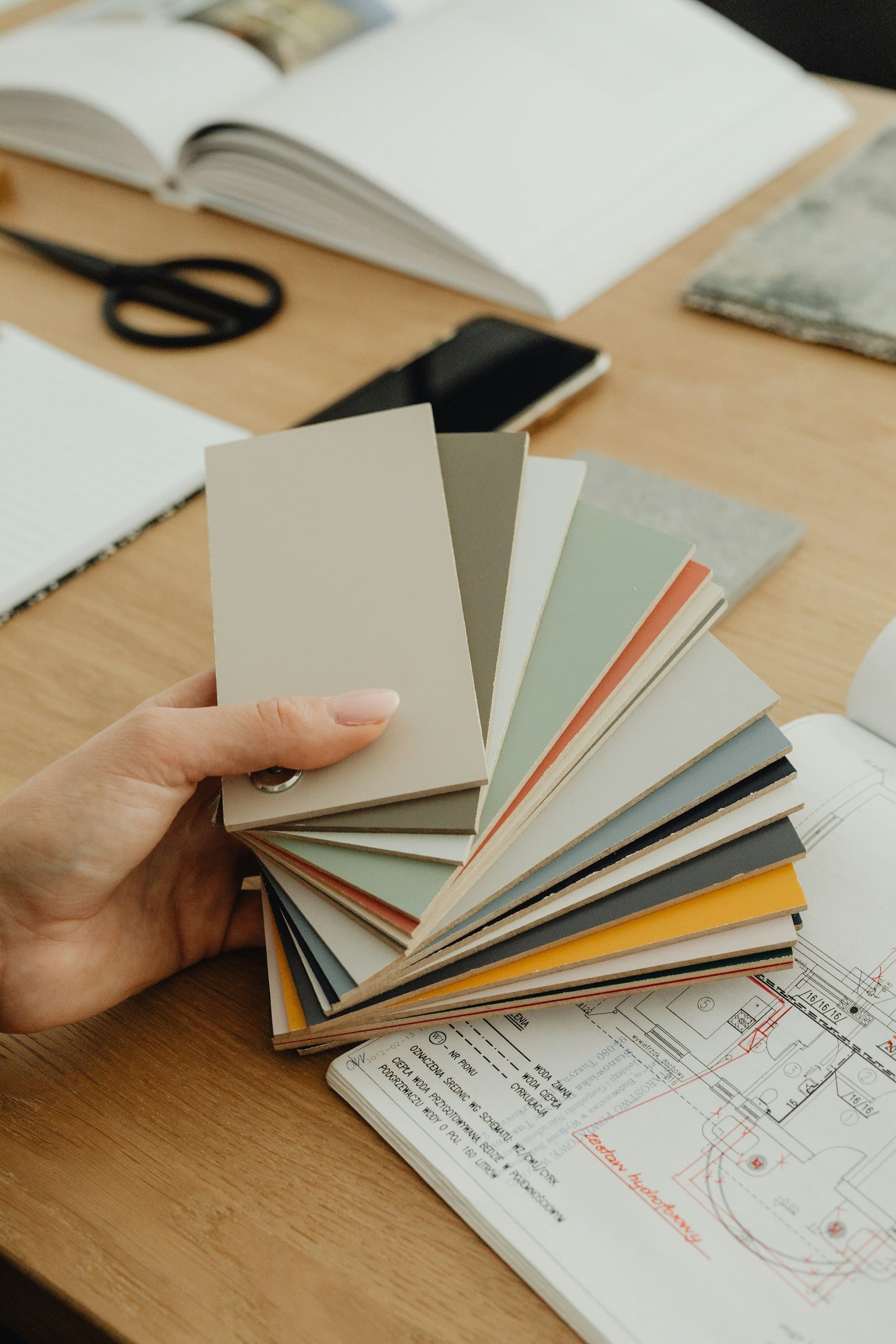 A hand holding a fan of muted color swatches above drawings, an open book and scissors on a desk