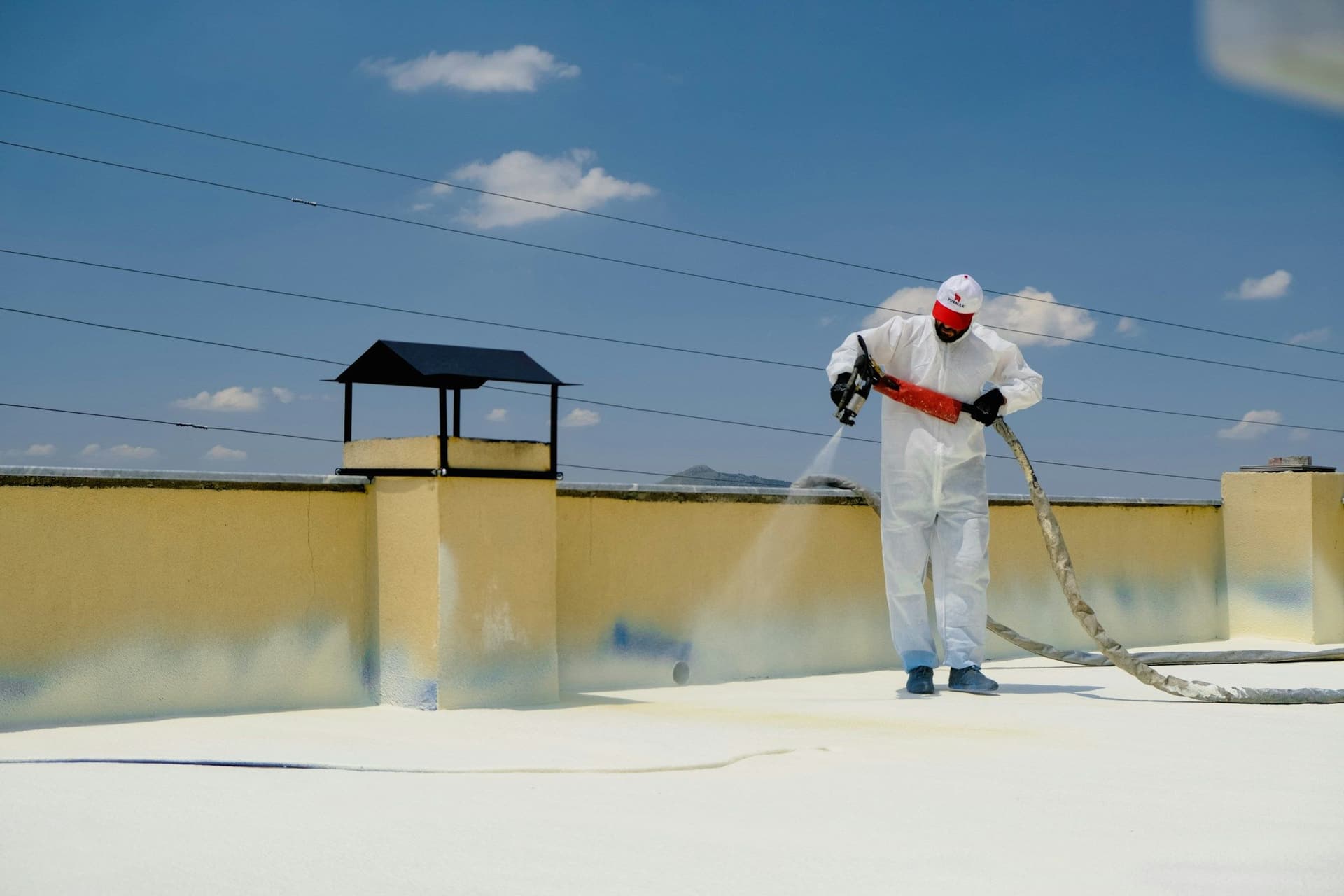 A worker in a white protective suit sprays a pale material over a flat roof beside a parapet wall