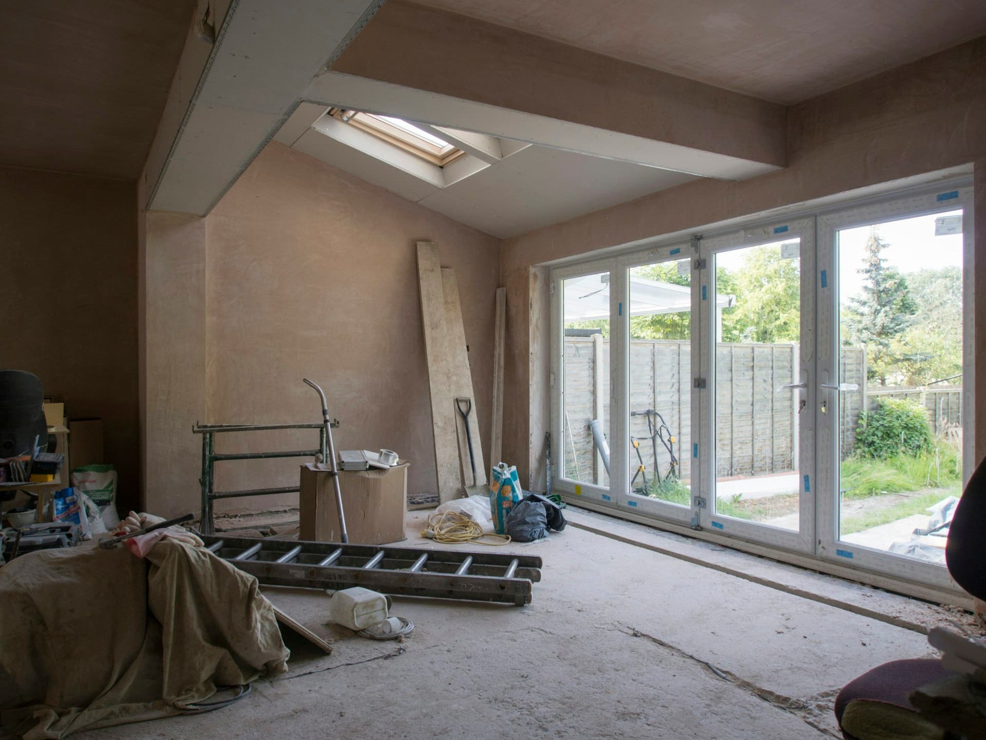 A new room mid-build with plastered walls, a skylight, glass doors to the garden, a ladder and tools