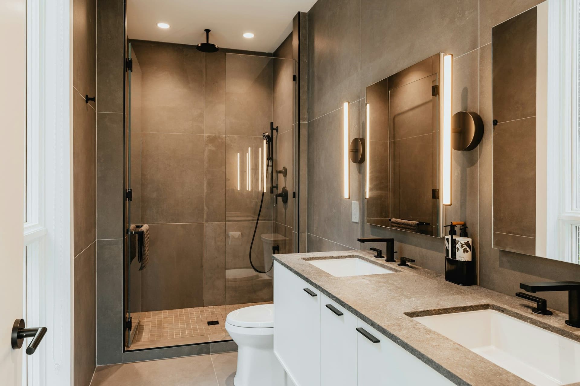 Bathroom with a glass shower in dark tile, a long white vanity with two basins and a toilet