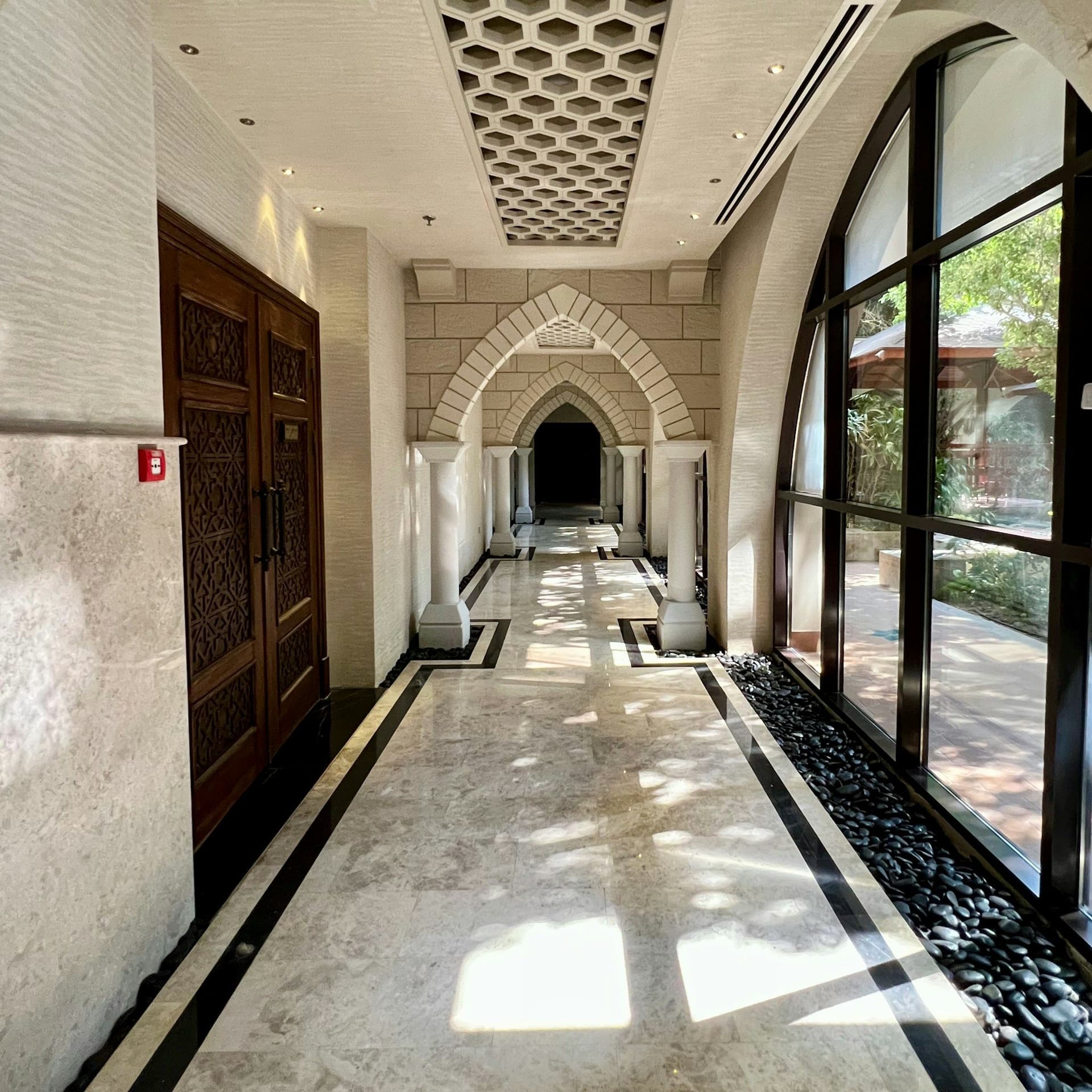Long hallway with a marble floor, carved wooden doors, large black-framed windows and a series of pointed arches