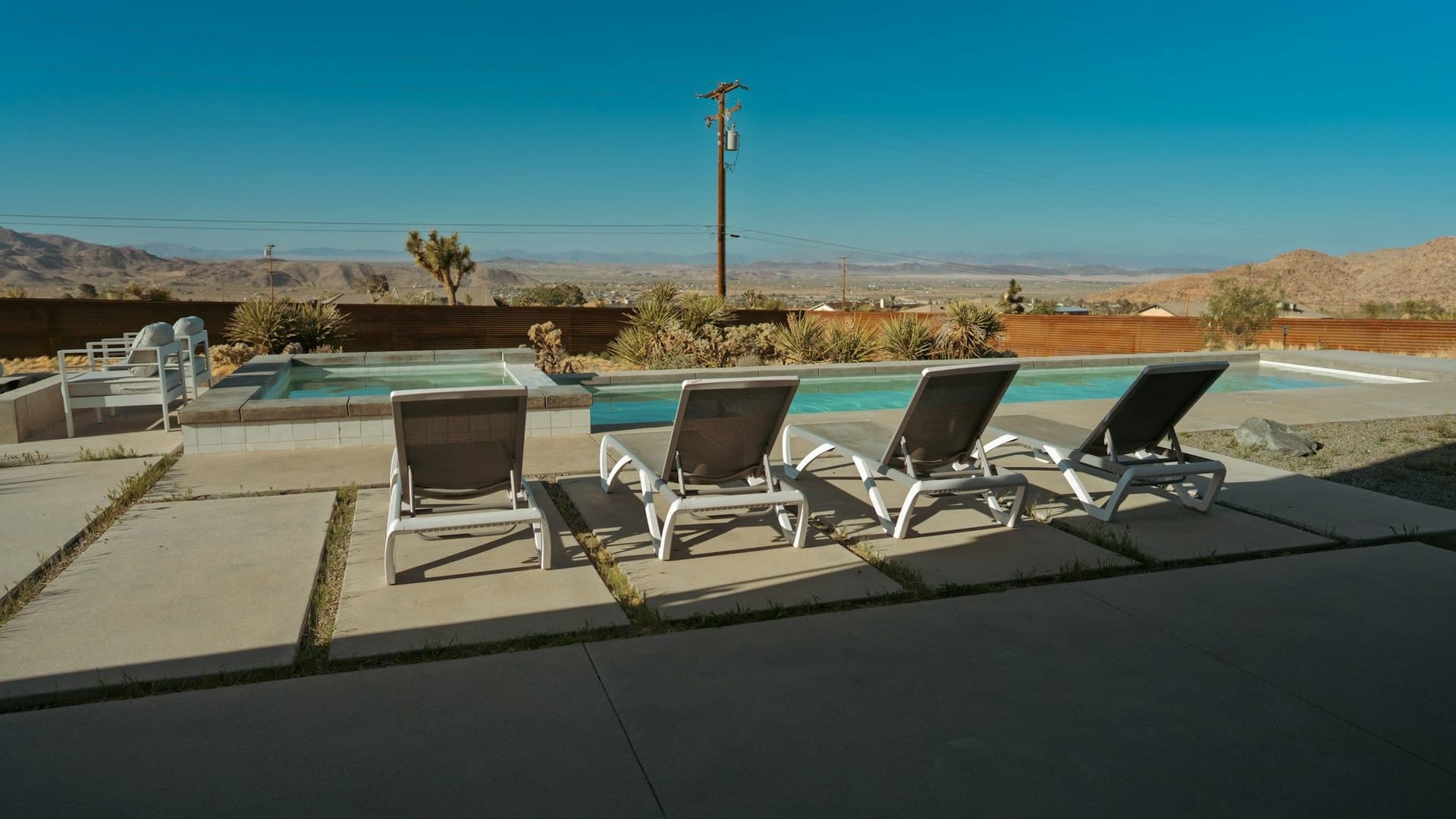 Row of lounge chairs on a concrete deck beside a pool with desert hills behind