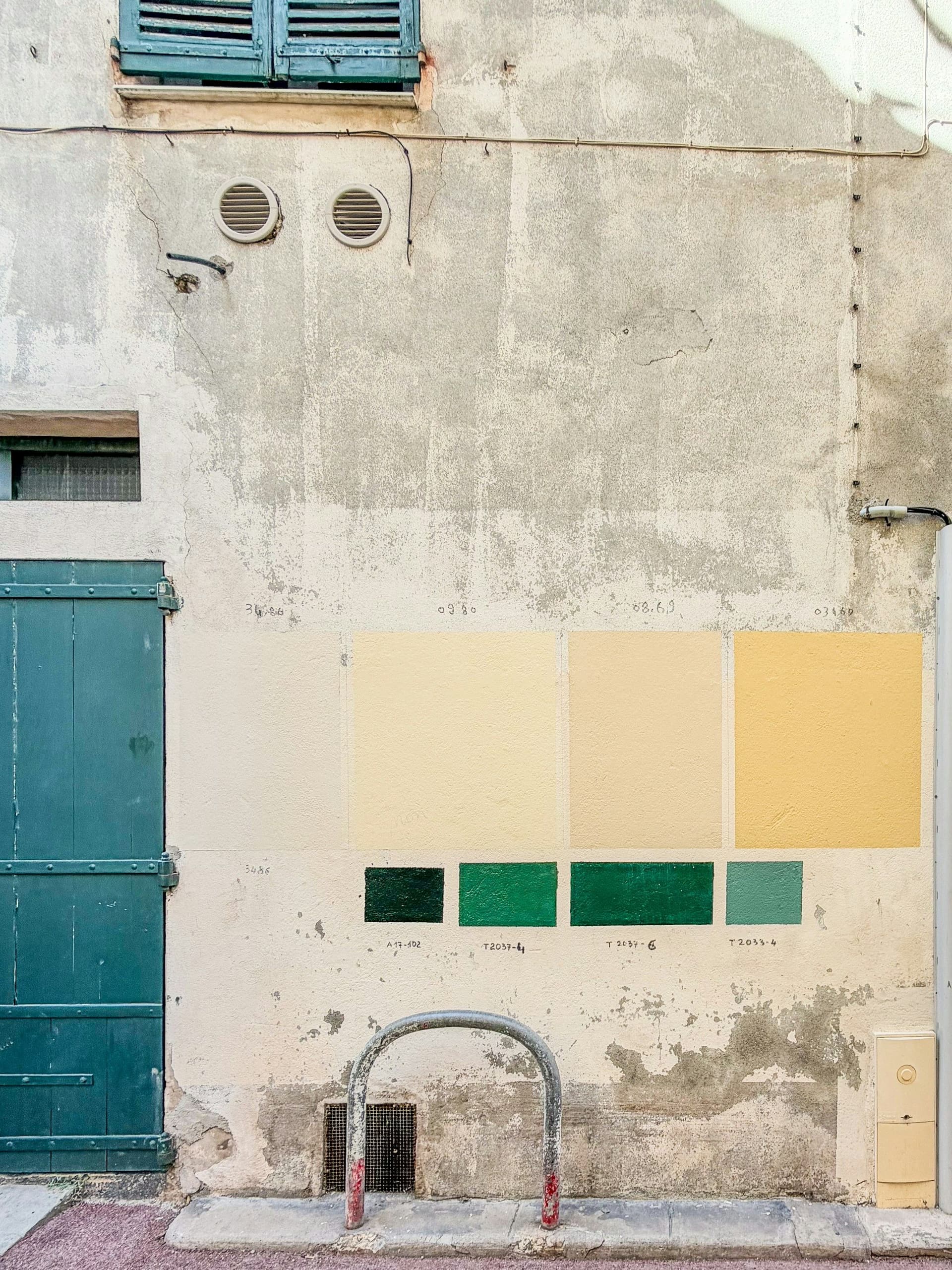 Square test patches of yellow and green paint on a weathered exterior wall beside a teal door