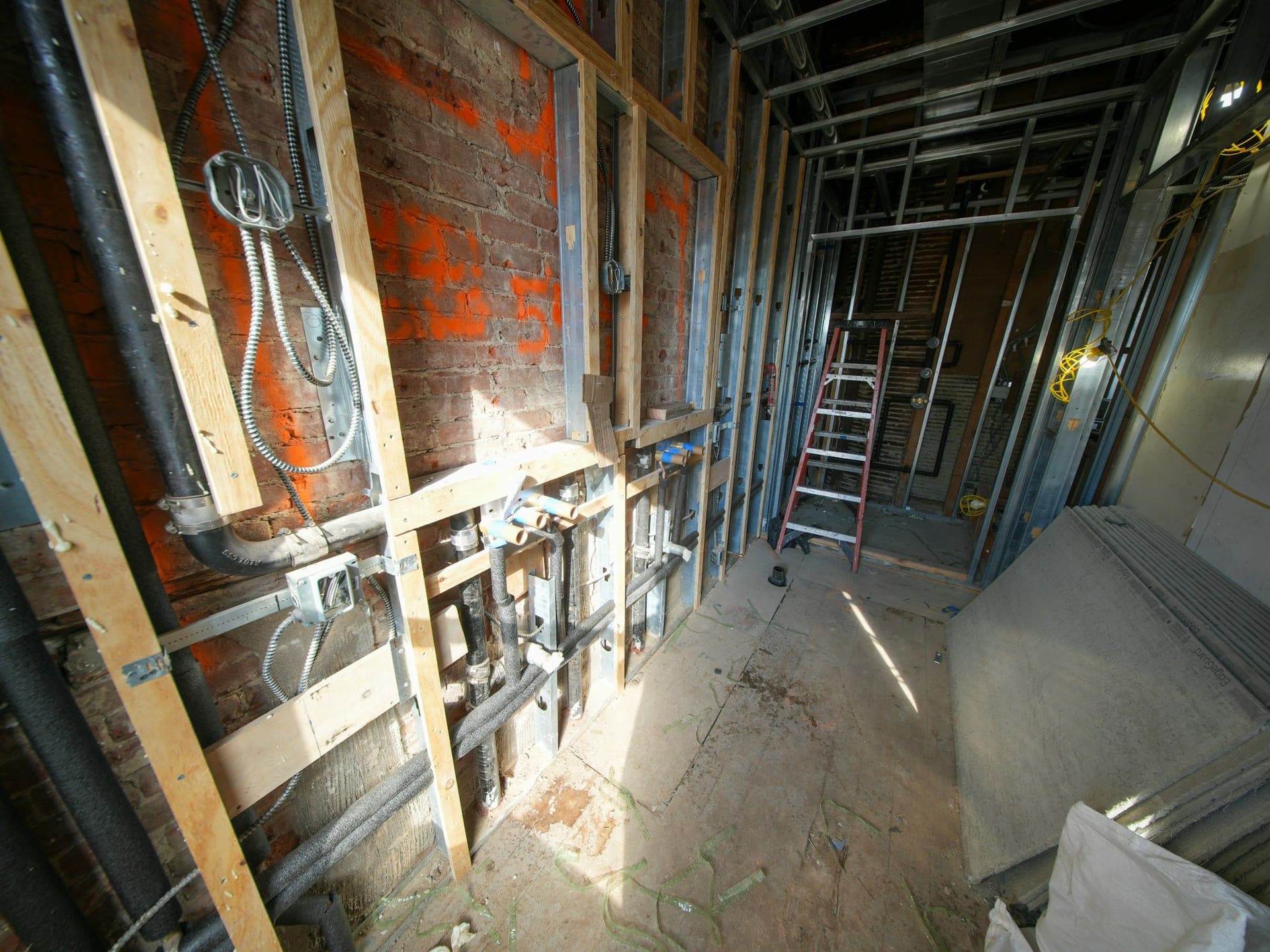 Unfinished room with exposed brick, electrical conduit boxes, pipes and wood framing, a ladder and drywall sheets on the floor