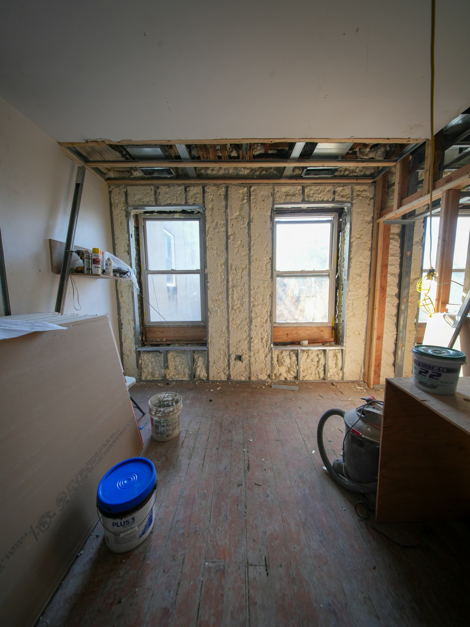 Room stripped to the studs, with drywall sheets leaning on one wall and a bucket on the bare floor