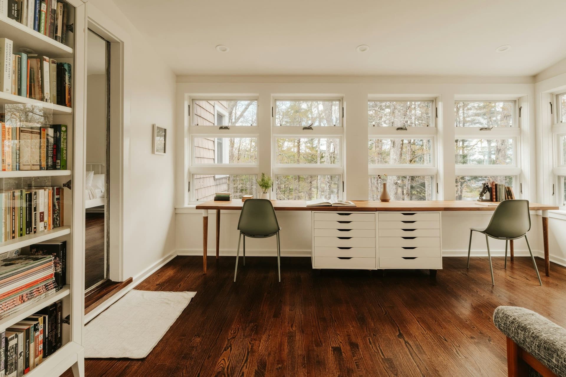 Bright room with a long wooden desk under a row of windows, two green chairs, white drawers and bookshelves