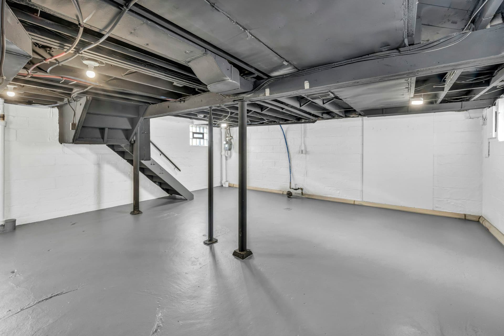 Unfinished basement with a painted concrete floor, exposed ceiling joists and ducts, steel support posts and a staircase