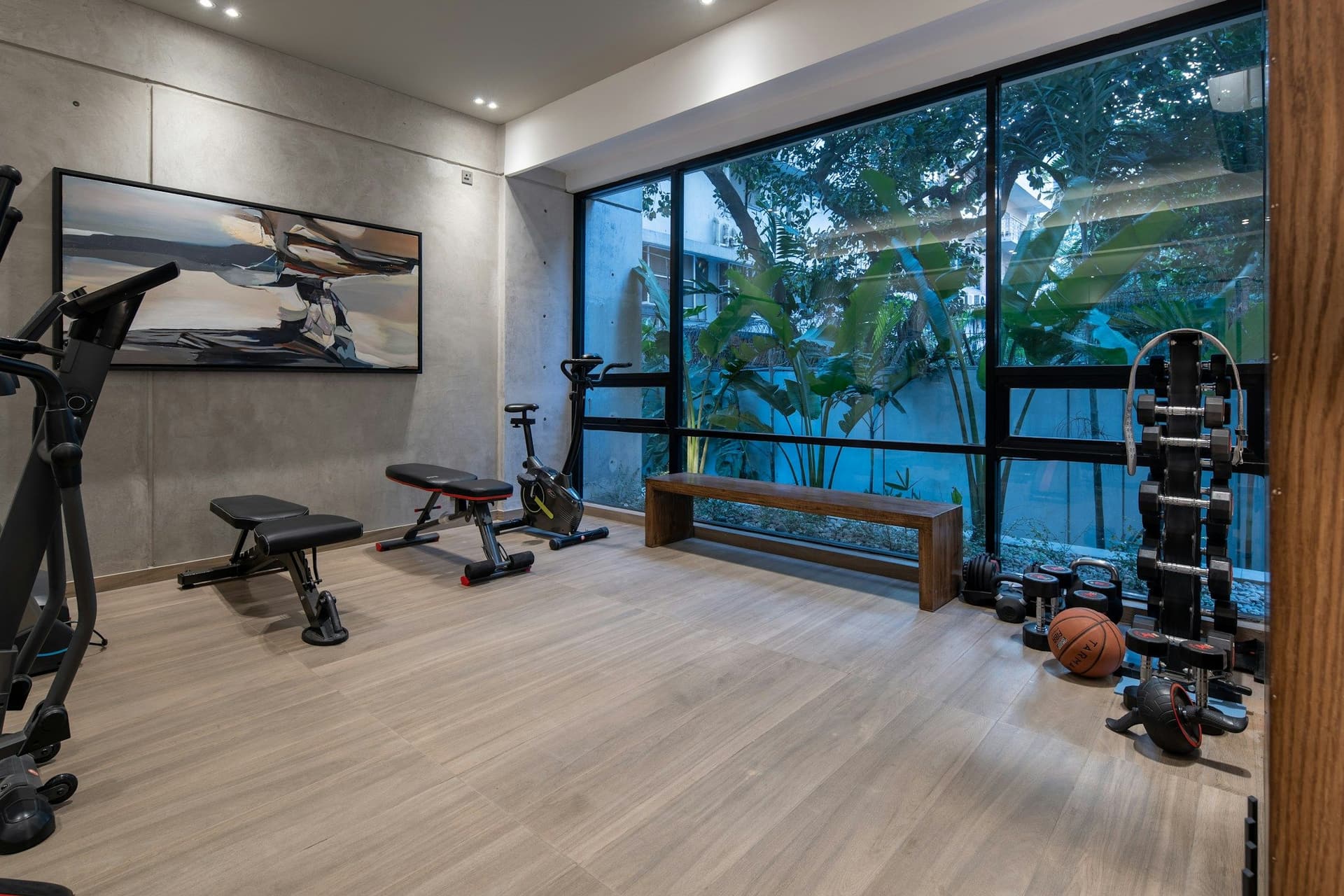 Bright home gym with a light wood floor, exercise equipment and a wall of black-framed glass looking onto greenery