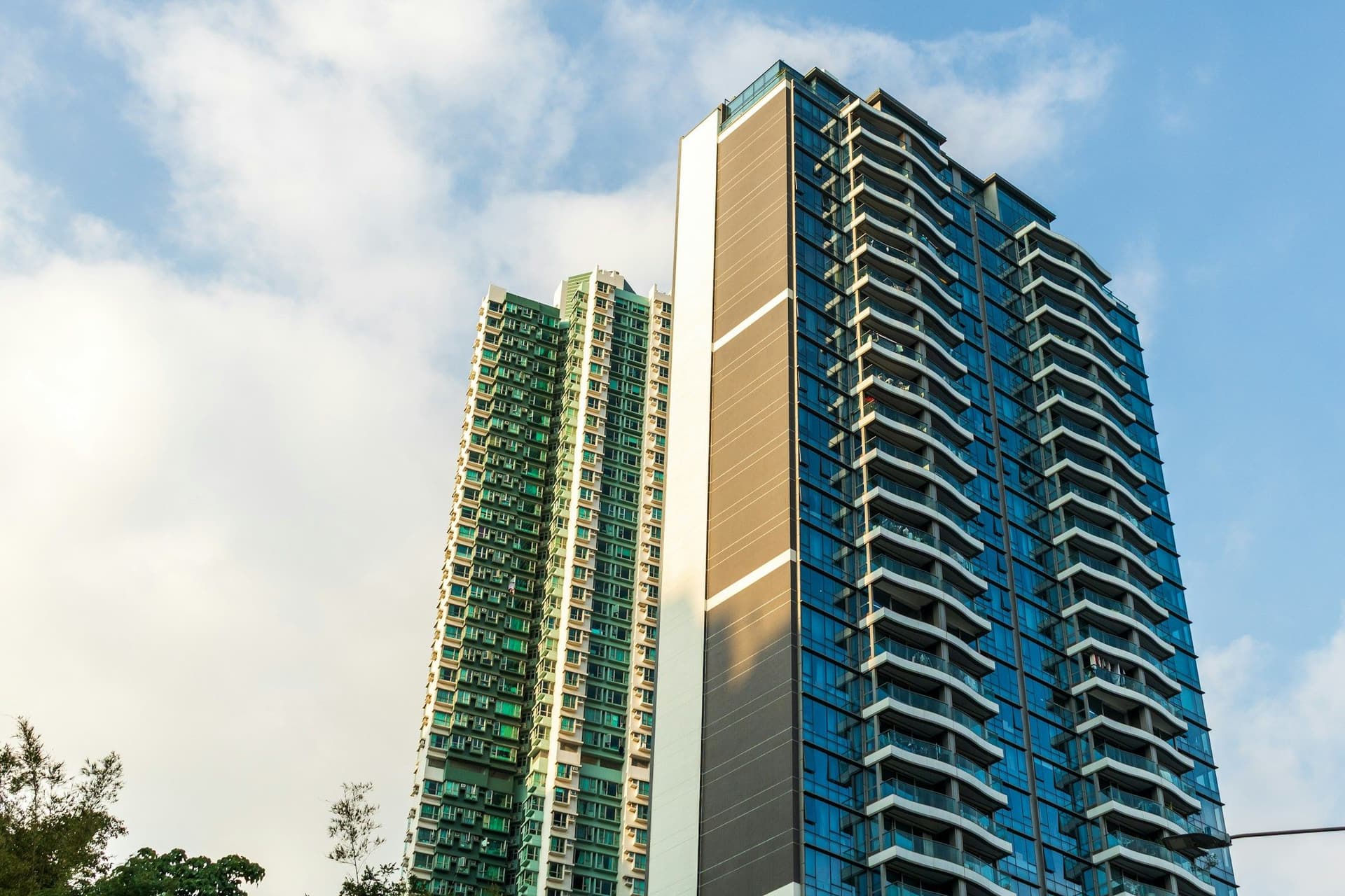 Two high-rise apartment buildings with glass balconies rising against a blue sky and white clouds