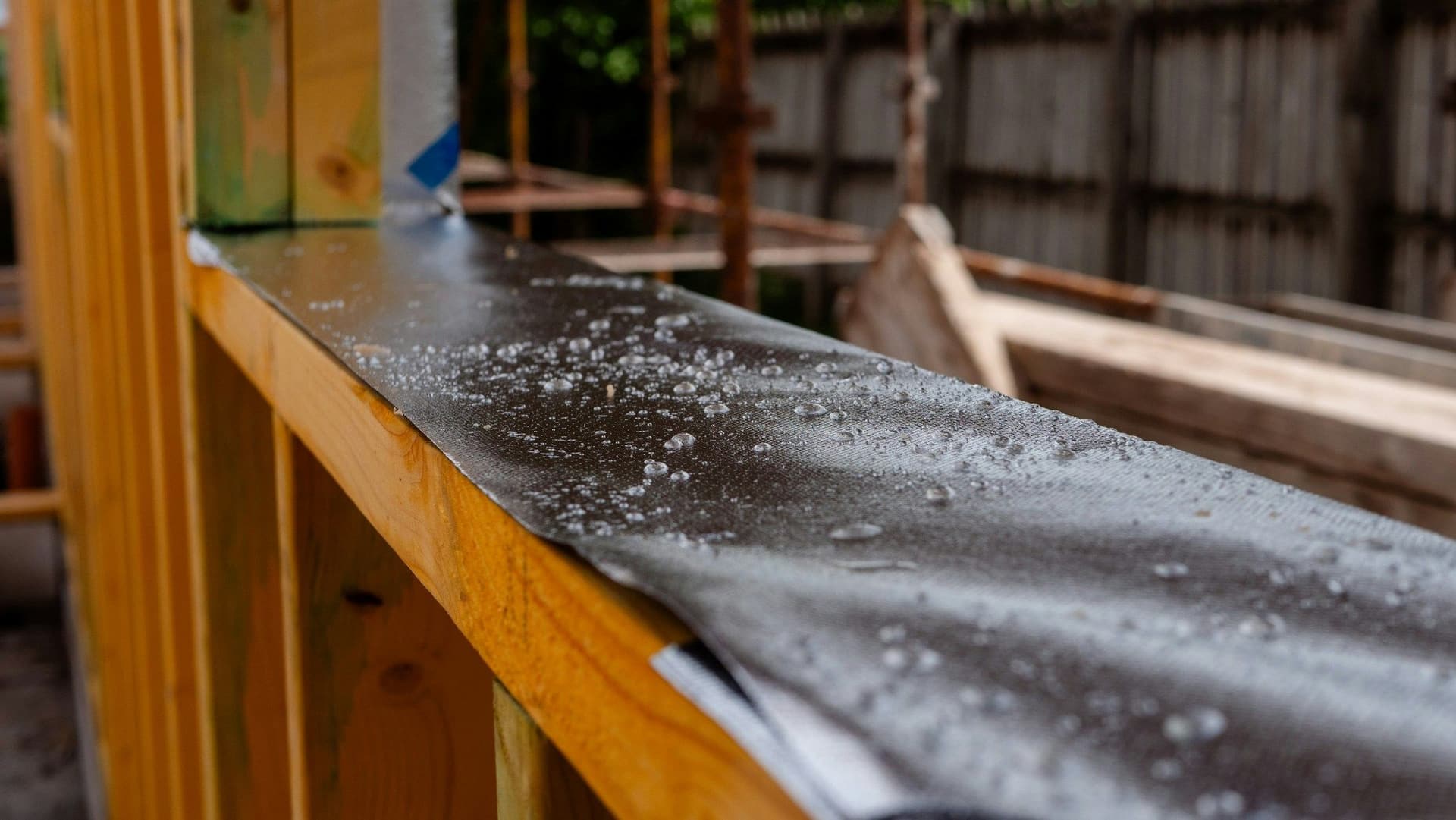 Raindrops bead on a black waterproof membrane draped over a timber wall frame