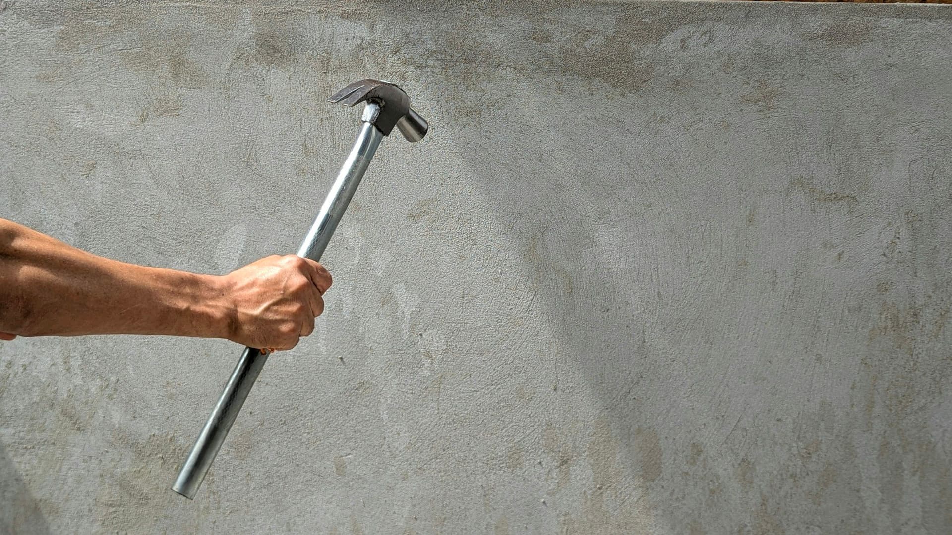 A raised hand gripping a claw hammer against a bare gray concrete wall