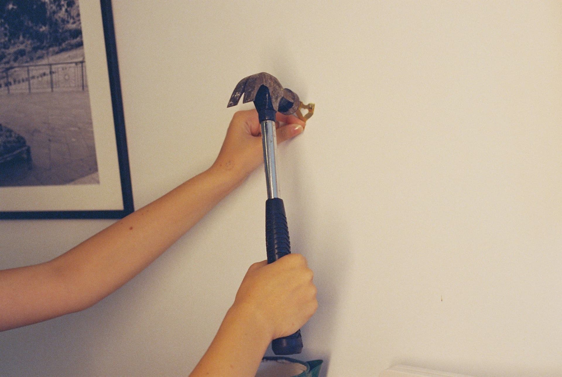 Two hands using a hammer to tap a picture hook into a plain wall beside a framed picture