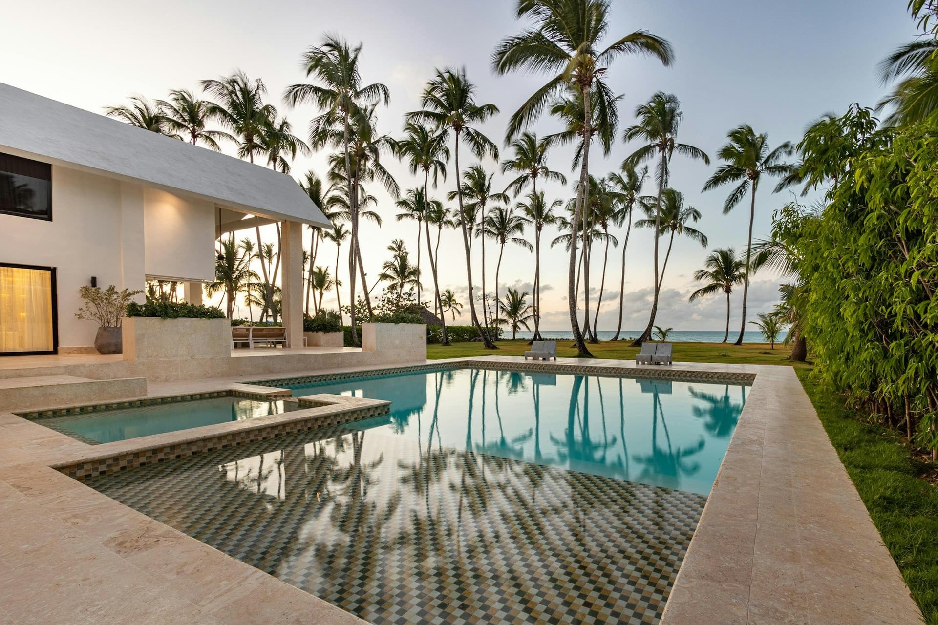 Mosaic-tiled swimming pool with travertine edges reflecting tall palm trees beside a modern white villa at dusk