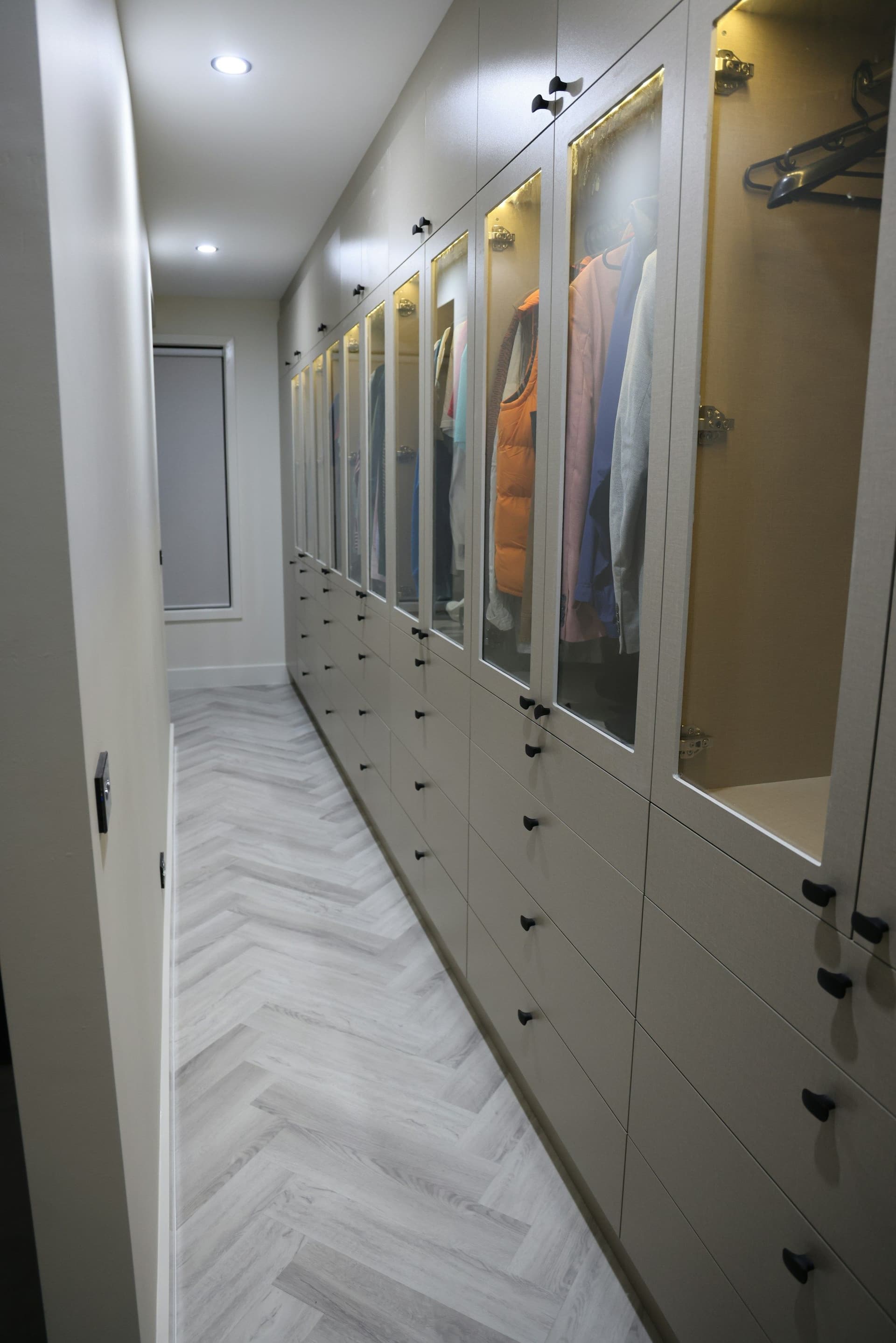 Long narrow walk-in closet with glass-fronted cabinets, drawers below and a herringbone floor