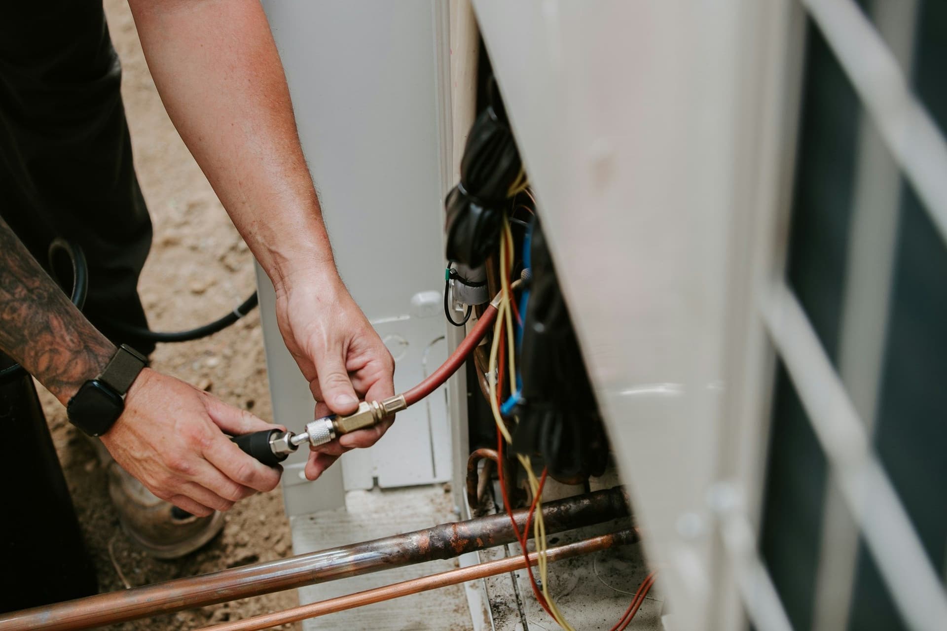 A technician's hands connect a hose to an outdoor air conditioning unit beside copper pipes